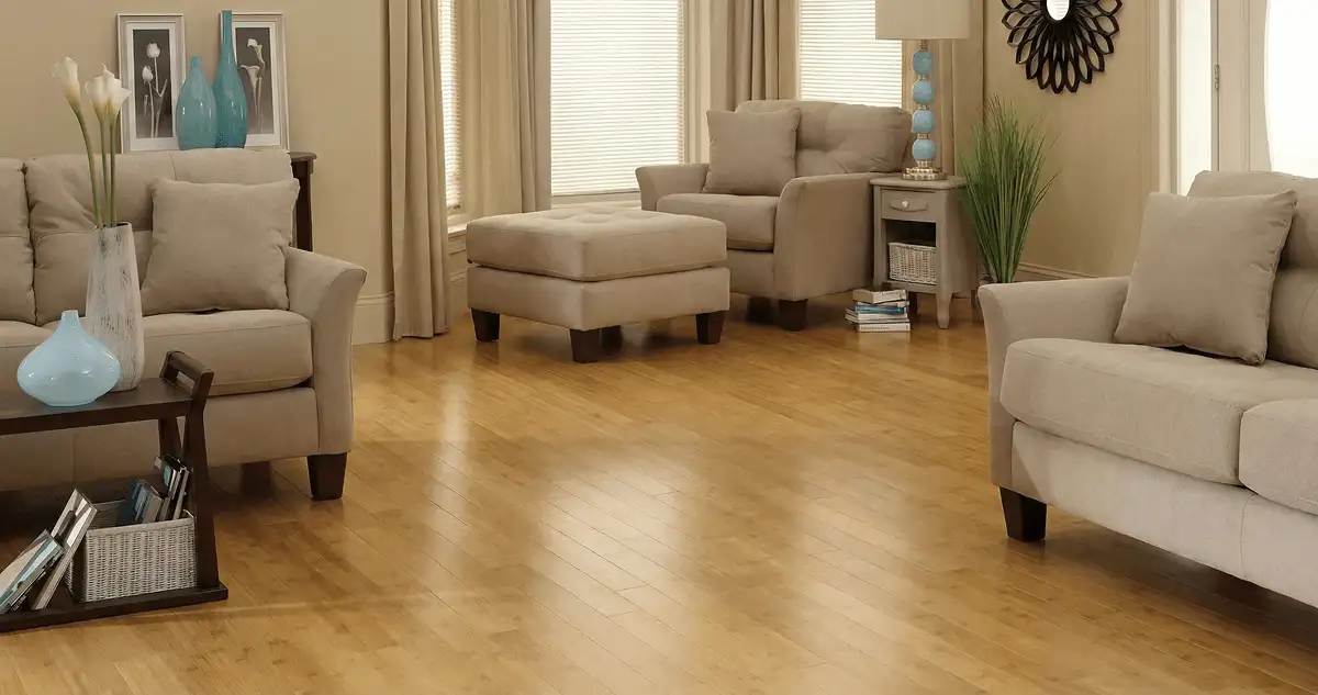 A living room with professionally installed bamboo flooring, beige sofas, a matching ottoman, a side table with a blue lamp, vases, framed pictures, and large windows with blinds and curtains in Wilmington, NC