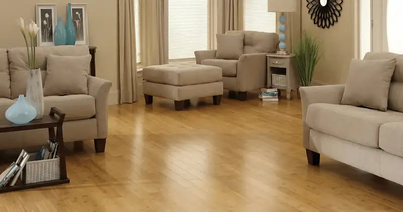 A living room with professionally installed bamboo flooring, beige sofas, a matching ottoman, a side table with a blue lamp, vases, framed pictures, and large windows with blinds and curtains in Wilmington, NC